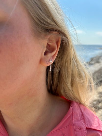 En kvinde med blondt hår står på en strand iført en koralfarvet skjorte. Hun bærer 'Sea Spark' øreringe fra VILMA, som består af en sølvfarvet søstjerne stud med små indlagte sten, og et hængende vedhæng med små gennemsigtige perler. Havet og himlen er i baggrunden, og der er sollys, der giver billedet en lys og sommerlig stemning.