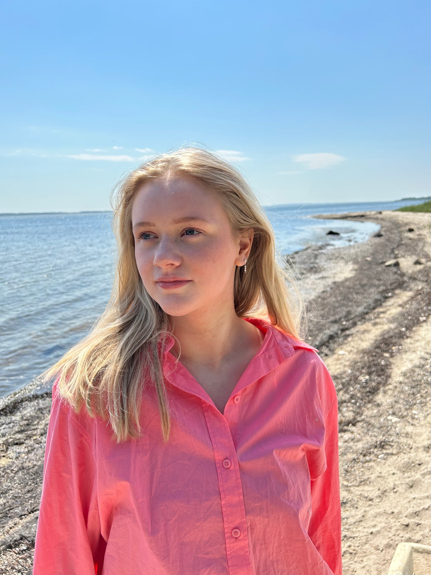 En kvinde med blondt hår står på en strand iført en koralfarvet skjorte. Hun bærer 'Sea Spark' øreringe fra VILMA, som består af en sølvfarvet søstjerne stud med små indlagte sten og et hængende vedhæng med små gennemsigtige perler. Havet og himlen er i baggrunden, og der er sollys, der giver billedet en lys og sommerlig stemning.