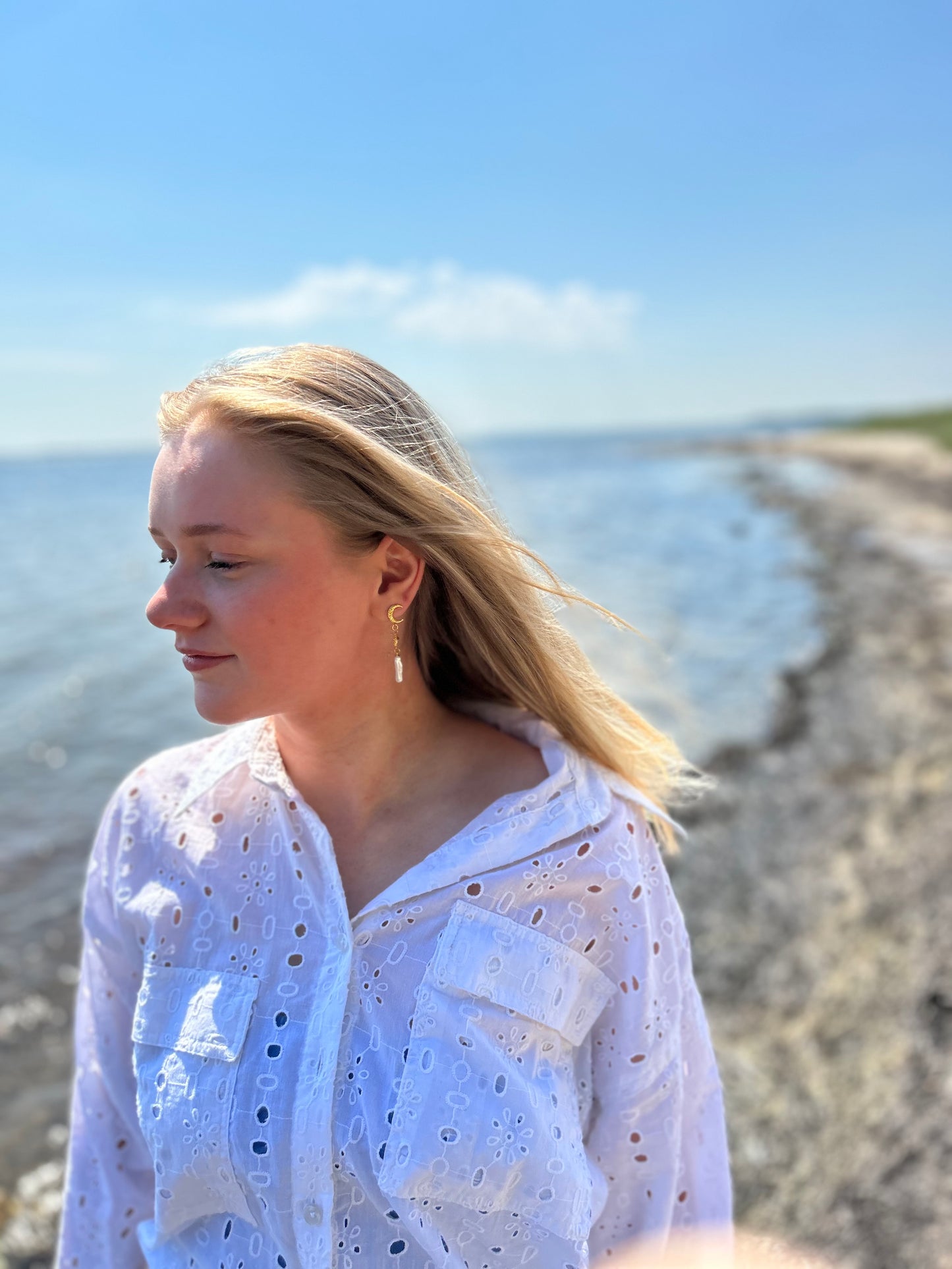 En kvinde med blondt hår står på en strand iført en hvid, hulmønstret skjorte. Hun bærer 'Lunar Glow' øreringene fra VILMA, som består af en guldfarvet halvmåne stud, et lille solvedhæng med en ædelsten i midten, og en lang, gennemsigtig dråbeformet perle. Havet og himlen er i baggrunden, og der er sollys, der giver billedet en lys og sommerlig stemning.