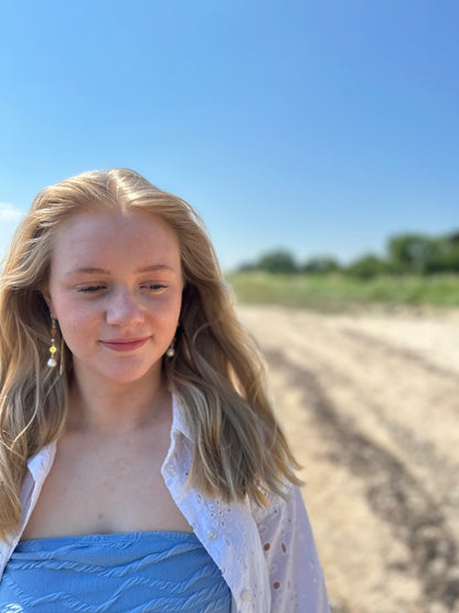 En kvinde med blondt hår står på en strand iført en blå stropløs top og en hvid, hulmønstret skjorte. Hun bærer 'Solar Drop' øreringe fra VILMA, som består af guldfarvede kroge, vedhæng med to små solformede dekorationer med ædelstene i midten, og en stor hvid perle nederst. Baggrunden viser en strand og grønt landskab under en klar blå himmel, hvilket giver billedet en lys og sommerlig stemning.