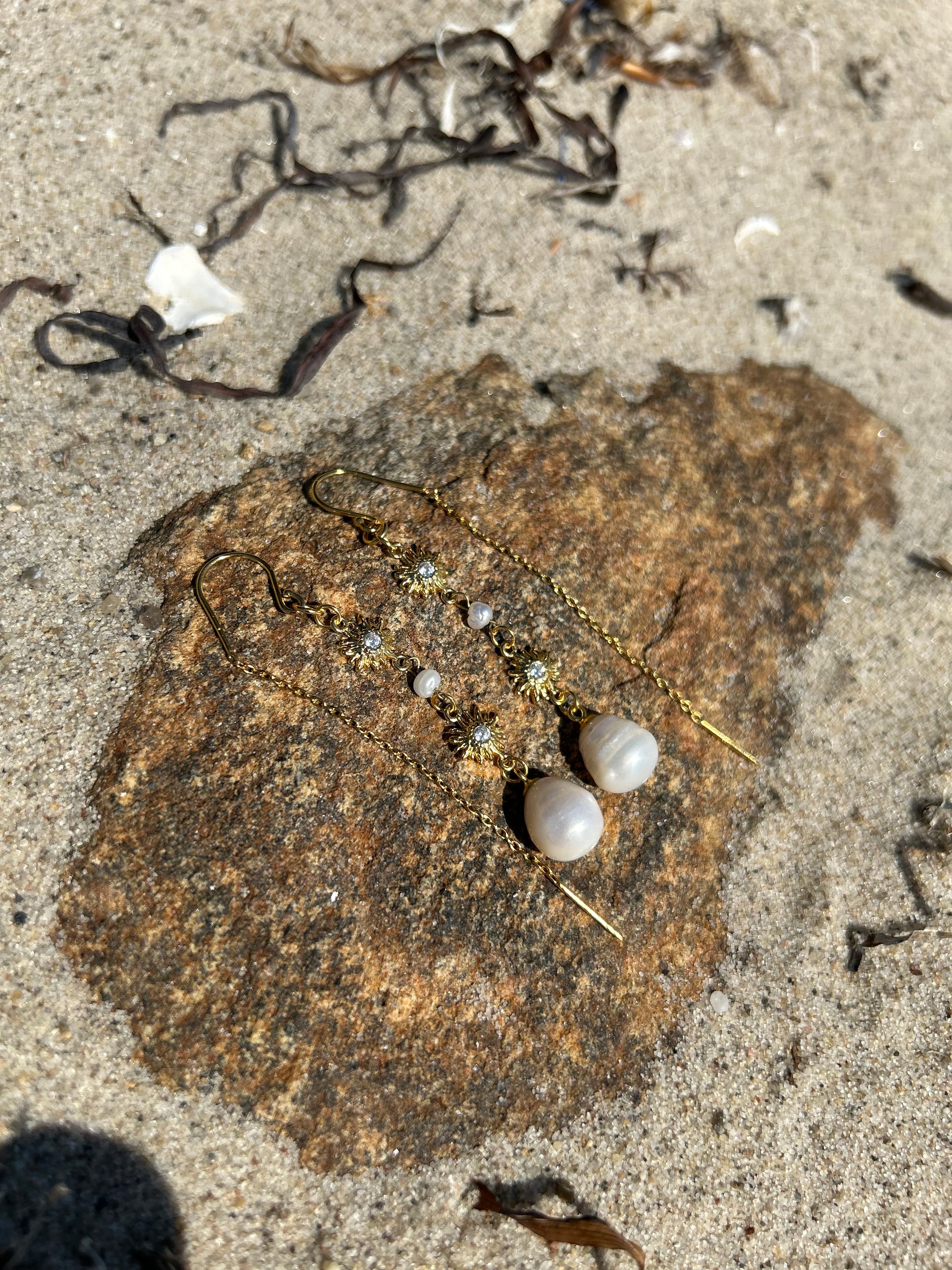 Et par 'Solar Drop' øreringe fra VILMA ligger på en klippeoverflade omgivet af sand og tang. Øreringene består af guldfarvede kroge, vedhæng med to små solformede dekorationer med ædelstene i midten, og en stor hvid perle nederst. Baggrunden viser et naturligt strandmiljø med sand og små stykker tang.